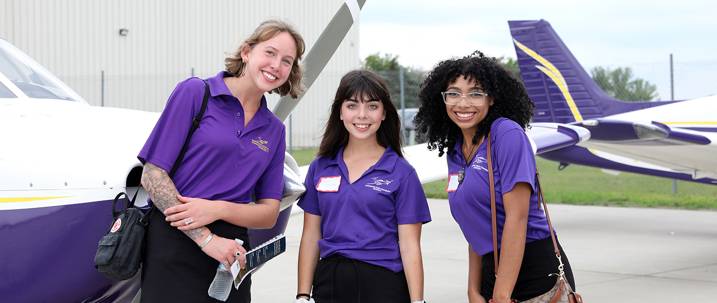 Three aviation students posing with smiles outside of the airport with the Maverick training airplanes behind them