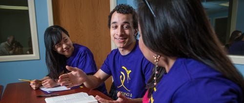 Three students sitting and talking at a table in a meeting room with a textbook, paper, pen and pencil