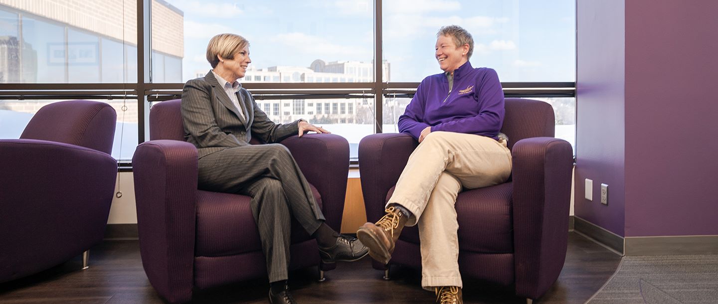 Two professors having a conversation while sitting on chairs by windows in a hallway on the Edina campus