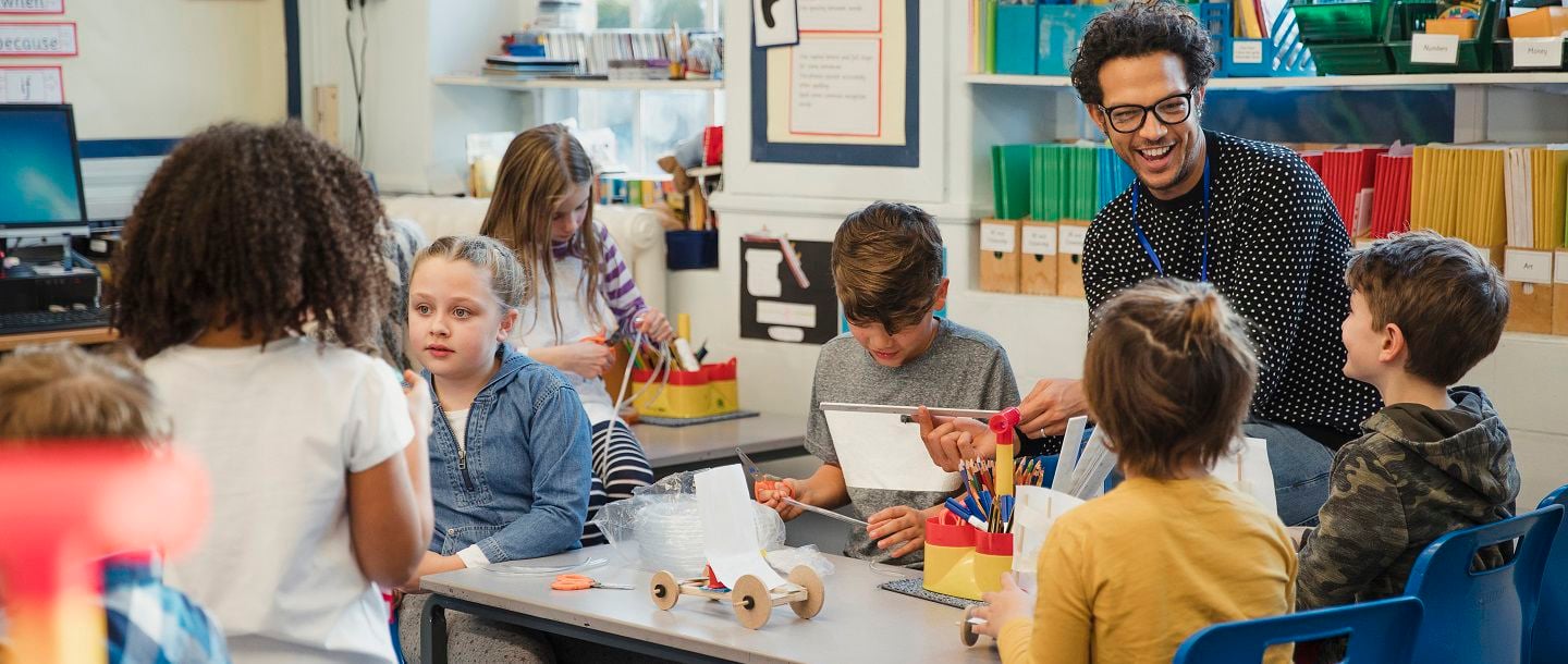 An elementary education student sitting at a table with four elementary students helping them with a project while other elementary students are working at different tables in the classroom