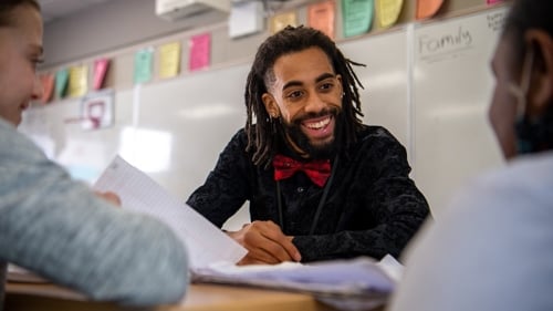 An elementary education student sitting at a table with a smile while helping two elementary students with their assignments in the classroom