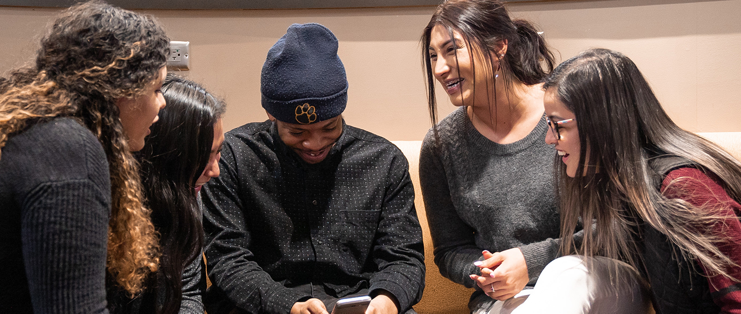 A group of students sitting down huddled together looking at cell phone and laughing