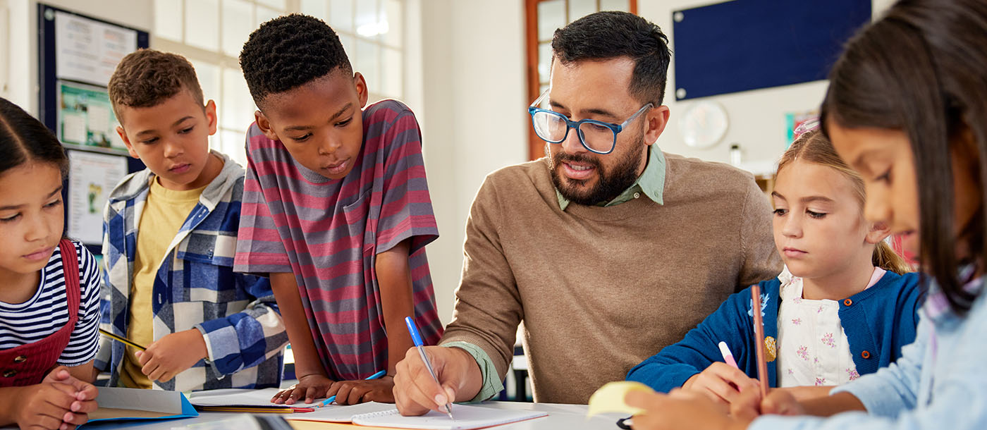 Diverse male teacher sitting at a desk with a few students around him watching him write on a paper. The kids are enthusiastic.